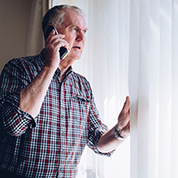 An older man looking out window of home with a phone in hand.